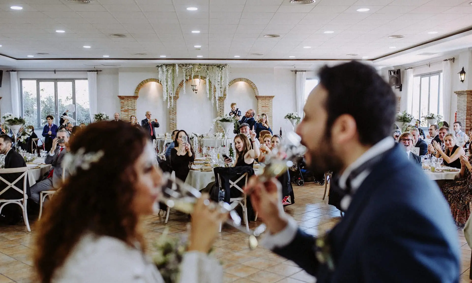 Los novios se dan de comer en el salón, lleno de invitados en plena fiesta