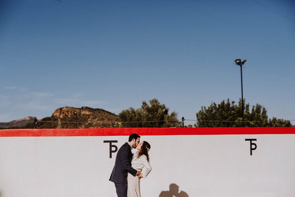 Una pareja de novios se besa ante la pared blanca de la plaza de toros, con el monograma TP de la finca, pinos y la roca de la sierra detrás